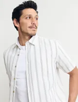 Camisa de manga corta a rayas verticales color natural y celeste, con cuello clásico, cierre frontal con botones de madera y bolsillo de parche en el pecho.