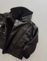 Campera tipo gamulán de cuero sintético negro con interior y cuello de corderito sintético al tono. Presenta cierre frontal, bolsillos laterales con solapa y broche metálico, y doble tira con hebilla en el cuello para ajuste.
