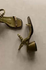 Sandalia de cuero color bronce metalizado, con punta cuadrada, taco ancho y bajo, y tiras finas adornadas con tachas esféricas plateadas.