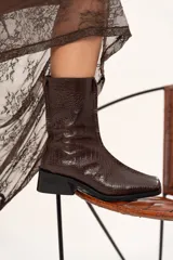 Bota de caña media con textura de reptil, confeccionada en cuero color chocolate. Presenta puntera cuadrada, taco bloque y cierre lateral interno.