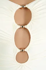 Malla enteriza de color crema con diseño strapless, fruncidos en el frente y tres aberturas circulares verticales separadas por herrajes metálicos brillantes. Presenta tirantes finos removibles.
