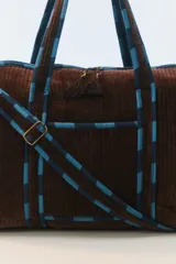Bolso de viaje tipo duffel de color gris oscuro, confeccionado en algodón acolchado con pespuntes verticales. Cuenta con asas de mano y una correa larga regulable con detalles de tiras en tonos fucsia y naranja, cierre superior y herrajes metálicos dorados.