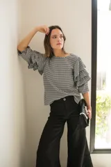 Blusa de tela con estampado vichy en blanco y negro, con cuello redondo y mangas con tres volados escalonados que llegan hasta el codo. Presenta puños elásticos y cierre con botón en la parte posterior.
