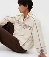 Campera de tejido técnico color beige con detalles de costuras en contraste color marrón. Presenta cuello alto, cierre frontal con cremallera, mangas largas con puños elásticos y estampa frontal con texto. Incluye un detalle bordado en la manga.
