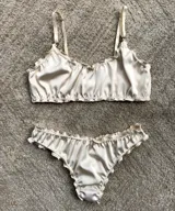 Conjunto de lencería compuesto por un corpiño tipo bralette y una bombacha colaless, confeccionados en satén color crema con terminaciones fruncidas. El corpiño cuenta con breteles regulables y doble capa de tela, mientras que la bombacha presenta un diseño de corte cavado con elásticos suaves.
