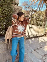 Sweater marrón de lana con cuello alto y manga larga. Presenta un copo de nieve blanco en el frente y bordados a mano. Tiene puños y cuello con diseño geométrico en tonos blanco, marrón y negro.