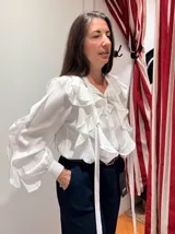 Camisa blanca de manga larga con volados frontales y lazos colgantes. Presenta mangas abullonadas con puños ajustados y cierre frontal con botones.