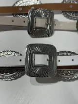 Cinturón marrón de cuero con hebilla plateada rectangular con diseño de flores y apliques metálicos circulares con diseño de flores.