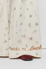 Pantalón de jean wide leg color beige con pequeños bordados y estampados coloridos dispersos, de tiro medio y corte amplio en las piernas.