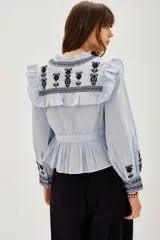 Camisa de algodón celeste con detalles de volados en los hombros y bordados en color negro en el pecho y las mangas. Presenta cierre frontal con botones y puños ajustados.