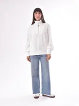 Buzo blanco de cuello alto con cierre frontal parcial y detalle de argolla dorada. Presenta un diseño de textura acanalada y mangas largas abullonadas con puños elásticos.