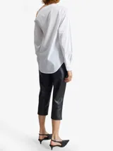 Camisa blanca de algodón con diseño asimétrico, escote cruzado con botones en diagonal, hombro descubierto con tirante fino, manga larga con puños abotonados y bolsillo de parche en el pecho.