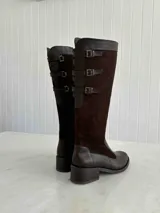 Bota alta de cuero color marrón chocolate, con taco cuadrado de 5 cm y caña alta hasta la rodilla. Presenta tres correas con hebillas en la parte superior de la caña. Fabricada en Colombia.