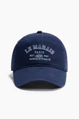 Gorra de sarga de algodón en color azul marino con efecto lavado, visera curva y diseño bordado en el frente con el texto 'Le Marais Paris Est. 1947 Maison Éternité'. Cuenta con botón superior y trabilla ajustable con cierre de metal en la parte trasera.