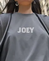 Remera oversize gris oscuro con corte holgado, mangas cortas y estampa trasera en blanco y negro con forma de estampilla que muestra a una persona con un gato y el texto 'Kaboa Industries'.