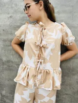 Blusa estampada con hojas verdes y blancas, con escote en V y mangas cortas abullonadas. Presenta volados en el ruedo y detalle de lazos en el frente.