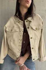 Sobrecamisa beige de gabardina con tachas metálicas decorativas en el cuello y la solapa frontal. Cuenta con bolsillos con solapa en el pecho y cierre frontal con botones.