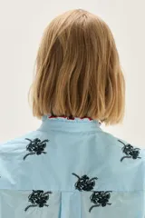 Blusa de algodón en color celeste con estampado floral en negro. Presenta cuello alto con volados y borde en color rojo, mangas abullonadas con puños elásticos tipo smock y cierre frontal con botones.