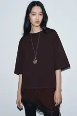 Camiseta de algodón de corte regular, color marrón oscuro o negro, con cuello redondo y manga corta. Presenta un bajo asimétrico con aplicación de encaje negro a tono.