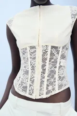 Top negro de cuello redondo y manga sisa, confeccionado con una combinación de tejido liso en el pecho y encaje transparente en el torso y los hombros. Cuenta con varillas metálicas forradas y cierre frontal con ganchos metálicos.