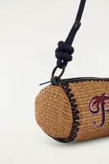 Bolso tipo bandolera de formato cilíndrico, confeccionado con material trenzado en color tostado, con detalle de bordado frontal que dice 'Rio' acompañado de una palmera y un tucán. Cuenta con una correa larga de cuerda oscura con nudos ajustables y cierre superior.