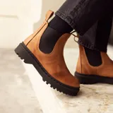 Bota tipo Chelsea de cuero color marrón, con paneles elásticos laterales en color negro, costuras a la vista y suela de goma dentada negra.