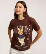 Remera de algodón color marrón oscuro con cuello redondo y manga corta. Presenta un estampado localizado con el texto "The Hamptons New York" y la ilustración de un conejo vestido de golfista.