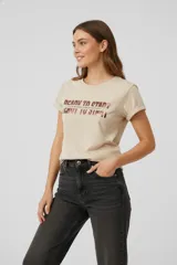 Remera de algodón color beige con cuello redondo y mangas cortas con dobladillo. Presenta un estampado frontal con texto en acabado metalizado tipo foil.