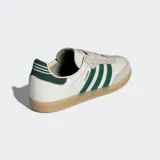 Championes Adidas modelo Samba OG, con diseño clásico de perfil bajo. Presentan una base de cuero color crema con superposiciones de gamuza gris en la puntera y los ojales. Incluyen las icónicas tres rayas laterales y el detalle en el talón en color verde oscuro, con suela de goma color caramelo.