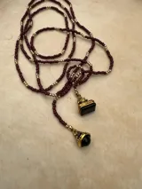 Collar largo de cuentas pequeñas en tono bordó, intercaladas con esferas doradas. Presenta dos dijes colgantes con piedras negras y detalles metálicos dorados. Su diseño permite usarlo largo o con doble vuelta.