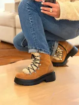 Bota tipo borcego de cuero marrón con suela track negra de goma. Presenta un aplique frontal desmontable de corderito sintético color crema y cordones negros con ganchos metálicos.