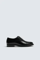 Zapato de vestir tipo oxford con puntera reforzada, confeccionado en piel lisa de color negro. Presenta cierre de cordones, forro interior y plantilla de piel, y suela con tacón bajo.