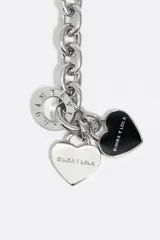 Pulsera de cadena metálica plateada combinada con perlas sintéticas irregulares, cierre tipo T y dijes colgantes en forma de corazón, uno de metal liso y otro esmaltado en negro con el logo de la marca.