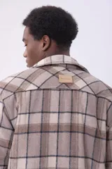Sobrecamisa de corte holgado con diseño a cuadros en tonos beige y marrón, confeccionada en tejido de textura suave. Presenta cuello clásico, cierre frontal con botones, dos bolsillos con solapa en el pecho y puños abotonados.