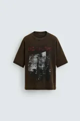 Camiseta de punto de algodón con corte boxy fit, cuello redondo y manga corta. Presenta un estampado frontal con efecto desgastado en tonos blanco y rojo sobre un fondo marrón oscuro/verde oliva lavado.