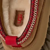 Pantuflas tipo mule UGG modelo Tazz II, confeccionadas en gamuza color tostado con plataforma alta. Presentan un detalle de costura trenzada decorativa en el borde superior en tonos rojo y beige, y el logo de la marca grabado en el lateral.