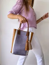 Bolso tipo tote de cuero color lila con textura croco, detalles en cuero marrón y laterales con franjas blancas. Incluye manijas de cuero marrón y accesorio de borlas colgantes.
