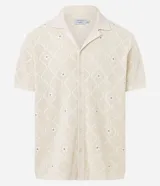 Camisa de manga corta color crema, confeccionada en tejido de punto con diseño de rombos calados y pequeños bordados florales en el centro de cada rombo. Posee cuello cubano y cierre frontal de botones.