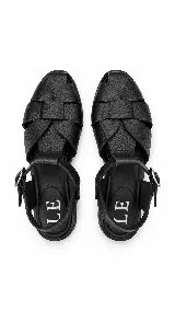Sandalias de cuero negro con diseño de tiras trenzadas artesanales en la parte delantera, plantilla de cuero y tira ajustable al tobillo con hebilla metálica.