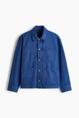 Sobrecamisa de lona de algodón en color azul, con cuello clásico, cierre frontal de botones y tres bolsillos de parche en la parte delantera. Presenta mangas largas con puños abotonados y canesú en la espalda.