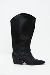 Bota de cuero negra de caña alta con polaina desmontable, taco cuadrado y punta fina. Presenta un arnés con argolla metálica alrededor del tobillo.