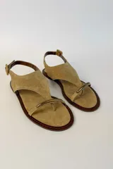 Sandalia plana de gamuza color tostado, con diseño de tira en T y argolla metálica en el empeine. Cuenta con tira ajustable con hebilla en el talón.