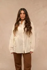 Camisa de manga larga color crema, con cierre frontal de botones, cuello camisero y detalles de cordones ajustables en los laterales de la cintura.