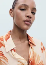 Camisa de corte recto y tejido fluido con estampado de palmeras en tonos naranja sobre fondo crema. Presenta cuello camisero, cierre frontal de botones ocultos, bolsillo en el pecho y mangas tres cuartos con vuelta.