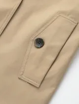 Gabardina corta de color beige, con diseño de doble botonadura, solapas anchas y cinturón ajustable en la cintura. Presenta bolsillos laterales y mangas largas.
