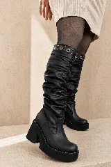Bota alta de cuero negro con caña fruncida, plataforma y taco cuadrado. Presenta detalles de ojalillos metálicos y tachas en el borde superior de la caña.