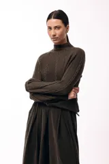 Polera de tejido de punto color marrón, con cuello alto y diseño de trenzas verticales. Presenta mangas largas con puños ajustados y un detalle de panel lateral asimétrico que se anuda en la cintura.