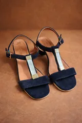 Sandalia de gamuza color azul marino con diseño de tira en T y aplique decorativo central de nácar. Posee pulsera al tobillo con hebilla ajustable y taco bajo cuadrado para mayor comodidad.