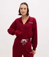 Campera deportiva tipo bomber de color bordó, con cierre frontal por cremallera, cuello redondo, mangas largas con puños elásticos y bolsillos laterales. Presenta un detalle de lettering bordado en el pecho.