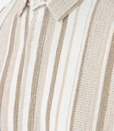 Camisa de hombre confeccionada en 100% algodón con textura, de manga corta y cuello inglés. Presenta un diseño de rayas verticales en tonos beige, blanco y marrón, con cierre frontal de botones.